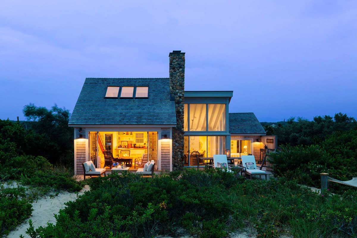 Cedar shingle beach cottage glowing at dusk with stone chimney, skylights, and open oceanfront deck