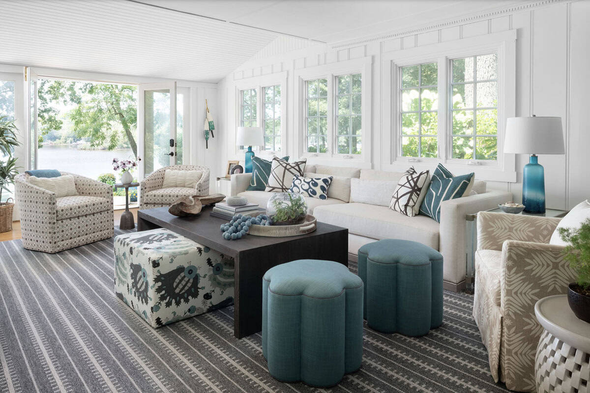 Bright coastal living room with teal ottomans, patterned armchairs, dark coffee table, and water views
