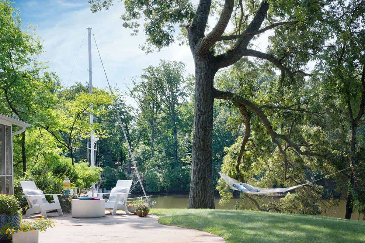 Waterfront patio with white Adirondack chairs, hammock, sailboat at dock, and lush tree canopy