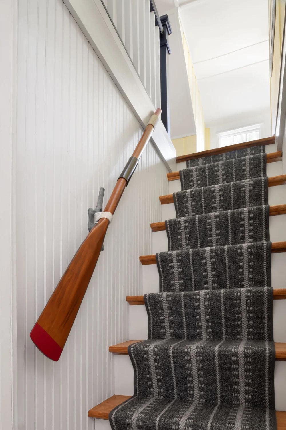 Staircase with wooden oar used as handrail accent against white beadboard wall and gray carpet runner