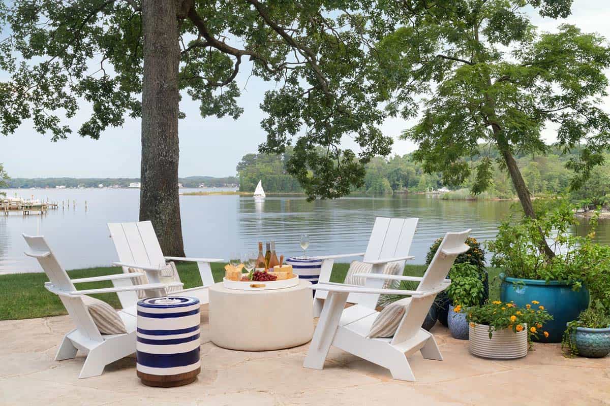 Waterfront patio with white Adirondack chairs, round concrete fire table, and sailboat on the river