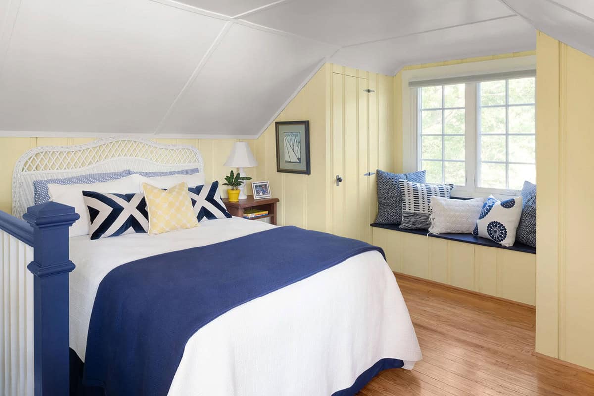 Attic bedroom with yellow beadboard walls, navy four-poster bed, white wicker headboard, and window seat