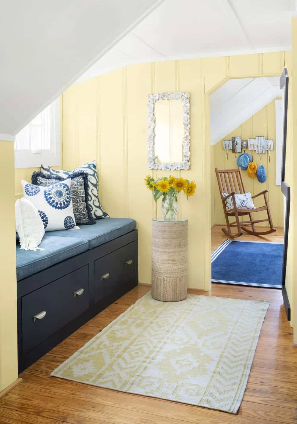 Yellow beadboard hallway nook with navy storage bench, sunflowers, shell mirror, and rocking chair beyond