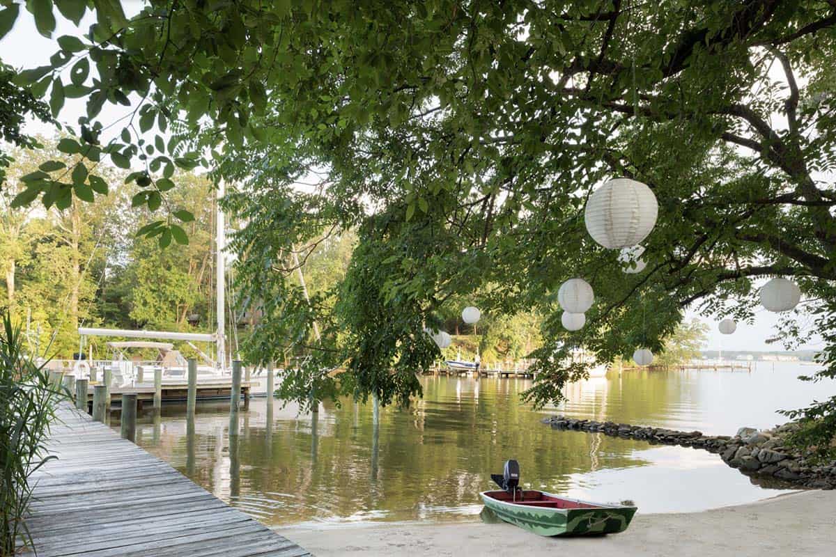 Private dock with paper lanterns hanging from tree canopy, sailboat, and small watermelon-painted motorboat