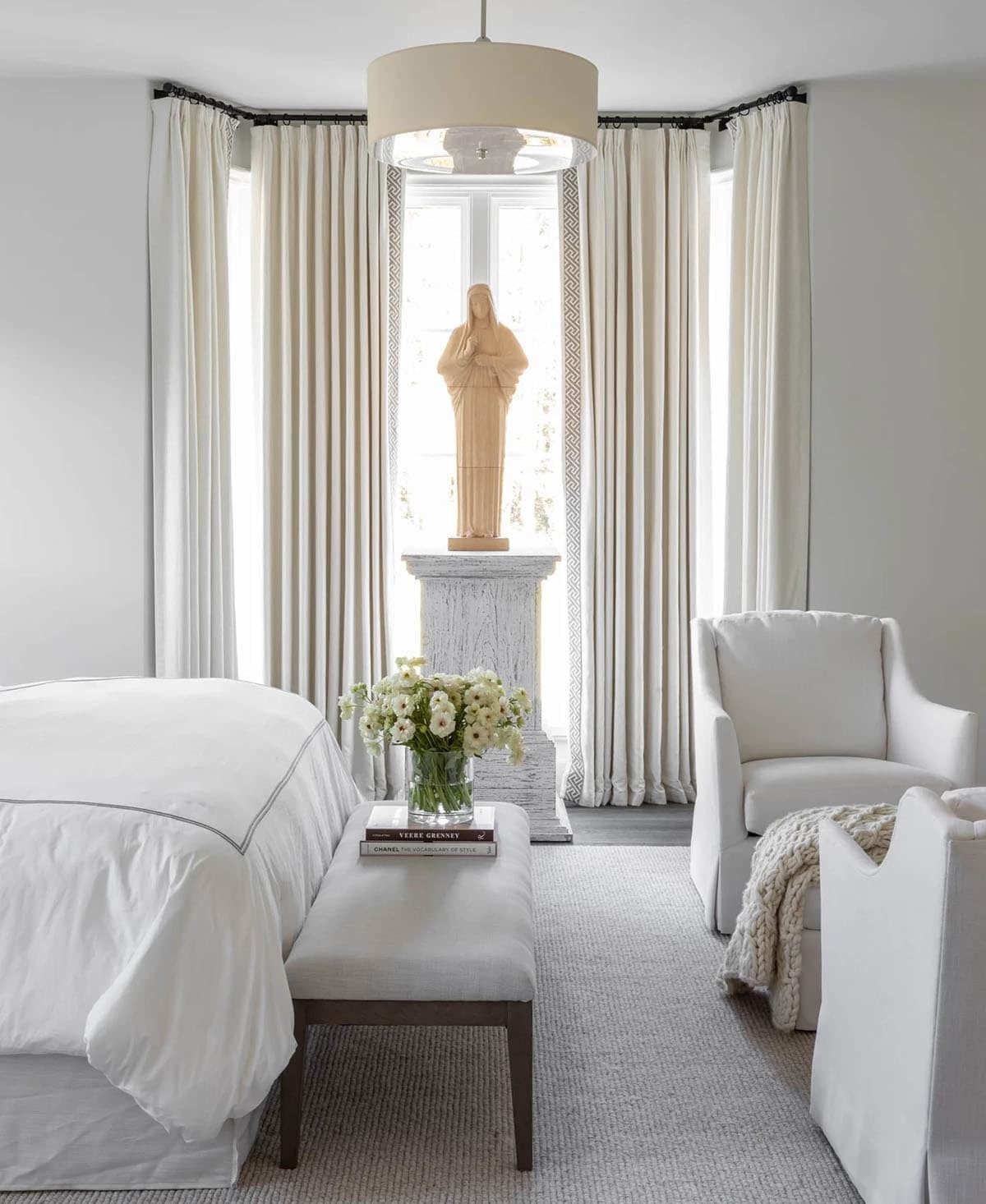 All-white bedroom with linen drapes, wooden religious statue on pedestal, and upholstered bench