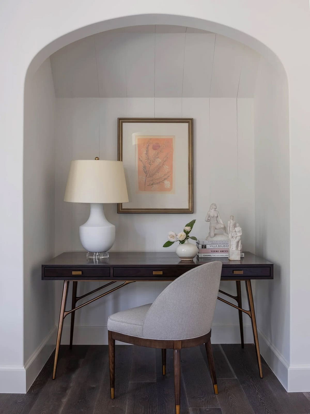Arched alcove home office nook with dark wood desk, white ceramic lamp, and botanical artwork