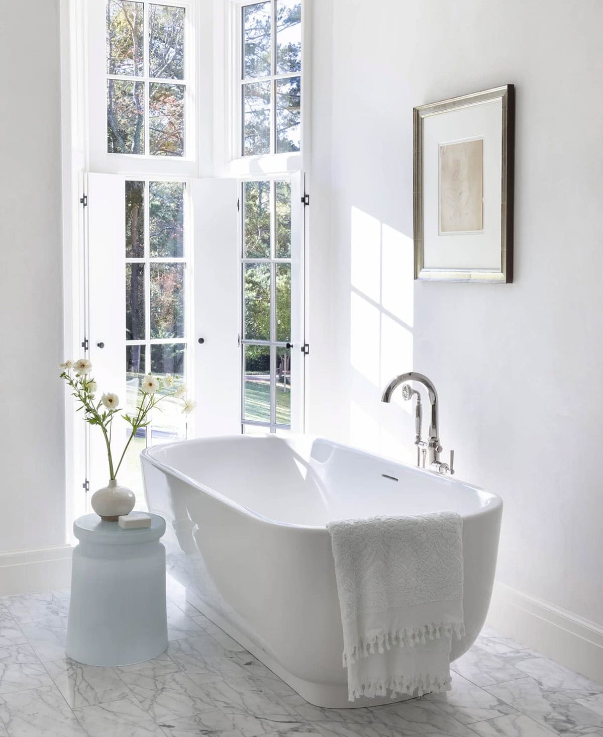 All-white primary bathroom with freestanding soaking tub, French doors, and marble tile floor