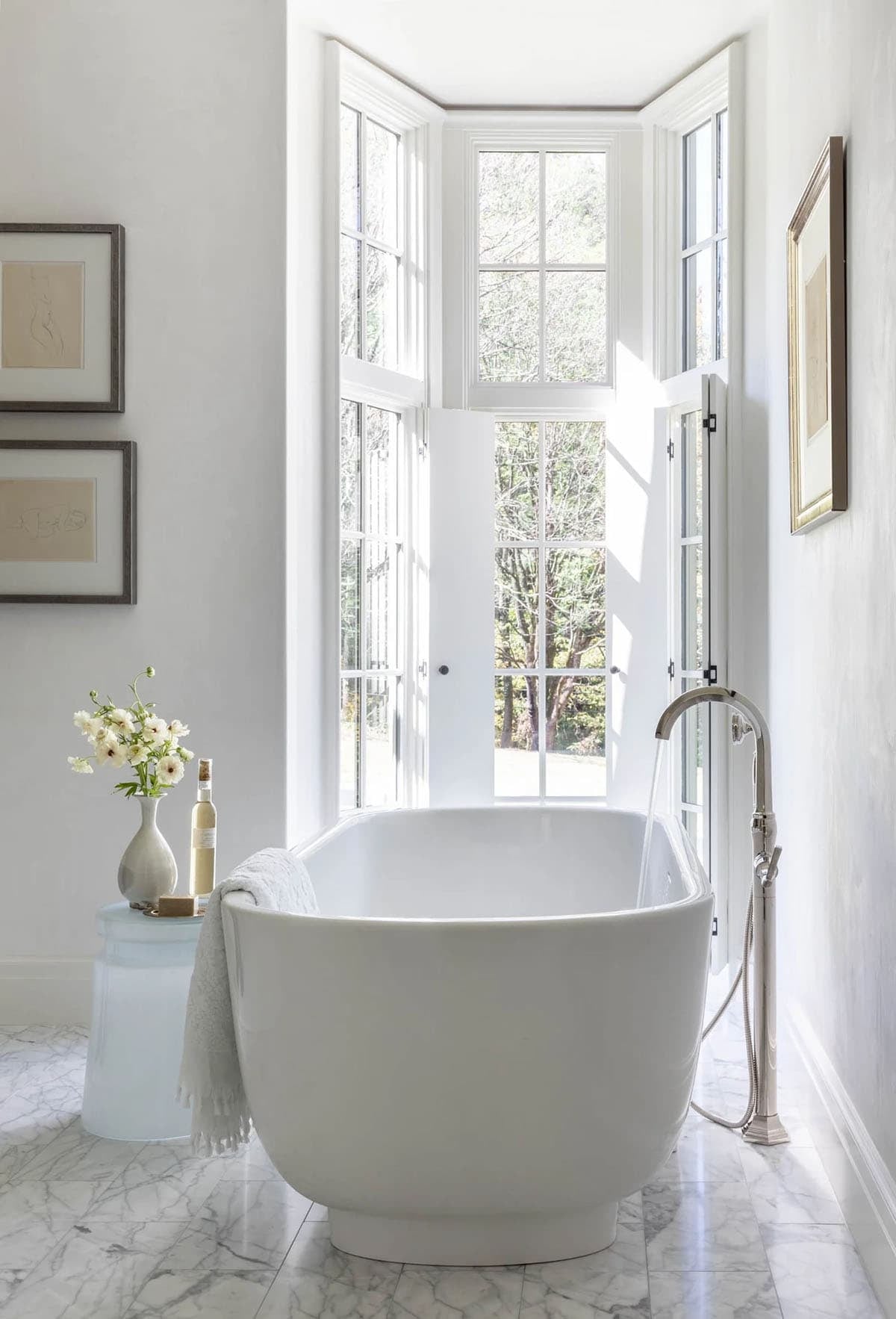 Freestanding soaking tub in bright white bathroom with bay window and framed figure drawings