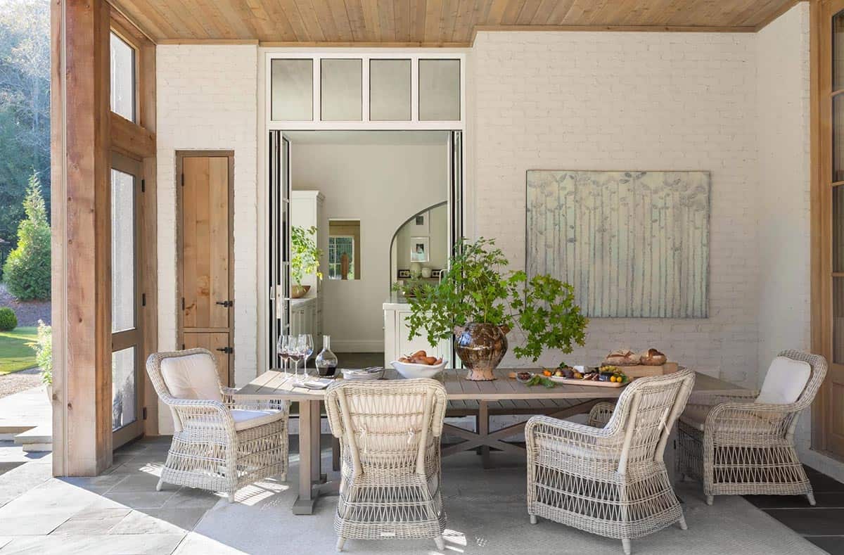 Covered porch dining area with wicker chairs, stone wall, and open view to kitchen interior