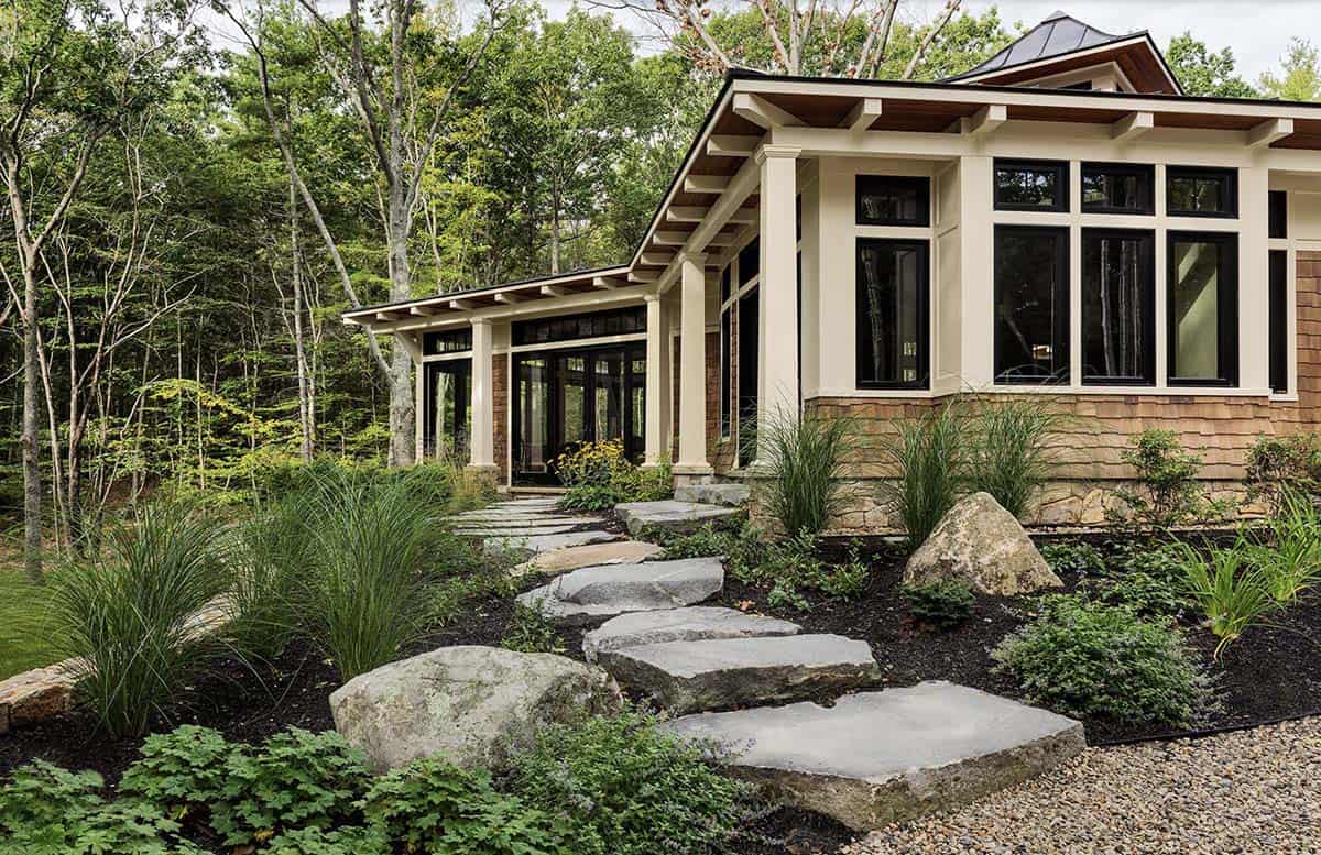 Stone step path leading to cedar shingle retreat with walls of black-framed windows amid Maine woodland