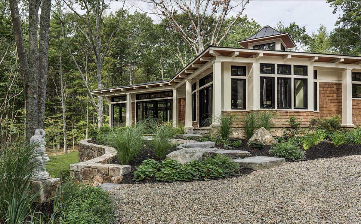 Rear view of cedar shingle retreat with stone retaining wall, gravel drive, and cupola above tree canopy