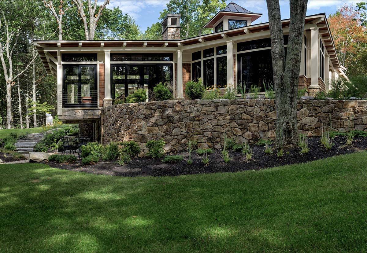Rear elevation of wooded Maine retreat with stone retaining wall, tiered landscaping, and wraparound glazing