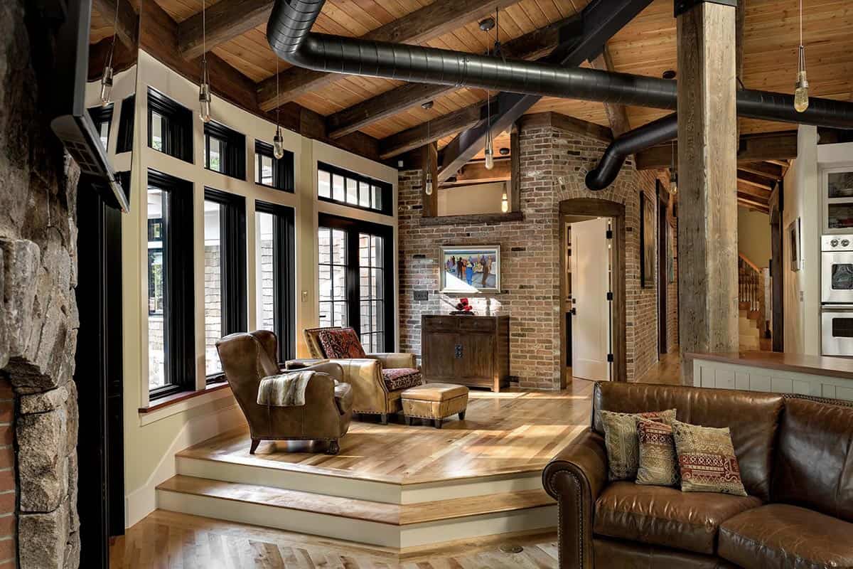 Cantilevered great room with stepped platform, leather armchairs, reclaimed brick walls, and black-framed woodland windows