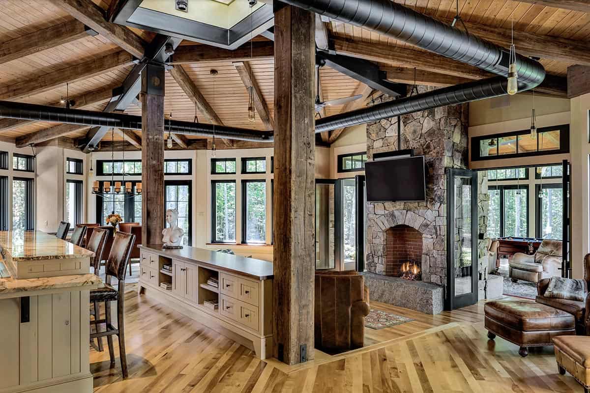 Open plan great room with floor-to-ceiling stone fireplace, dual-facing hearth, reclaimed timber posts, and woodland views