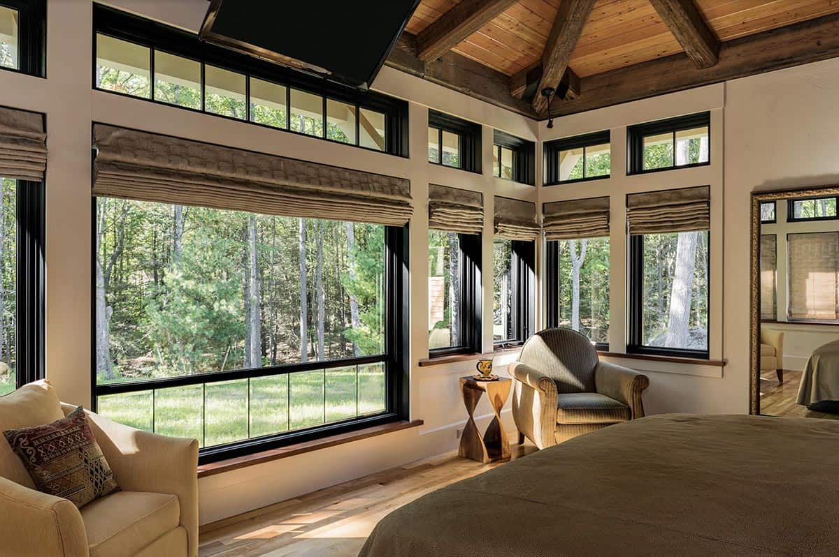 Main bedroom with wraparound wall of black-framed windows, timber beam ceiling, and sweeping woodland views