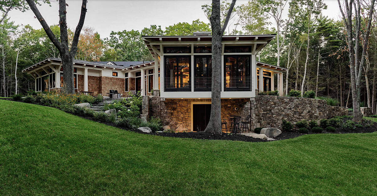 Rear elevation at dusk showing glowing interiors, cantilevered great room, stone retaining wall, and lower patio seating