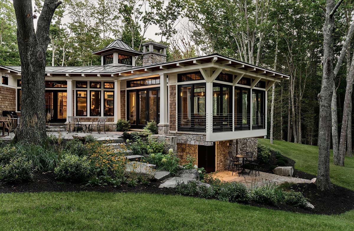 Evening exterior showing cedar shingle retreat with cupola, stone chimney, glowing great room pavilion, and tiered landscaping