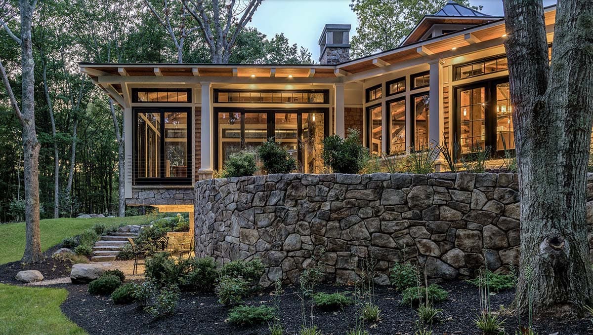 Twilight view of stone retaining wall, tiered garden plantings, and illuminated wraparound windows amid Maine woodland