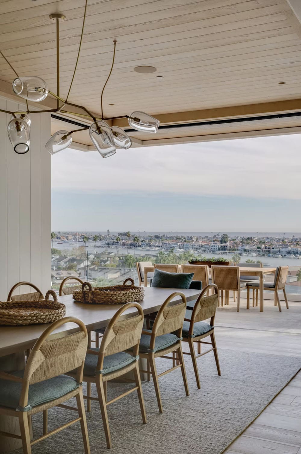 Indoor-outdoor dining space with rattan chandelier, woven chairs, and harbor views