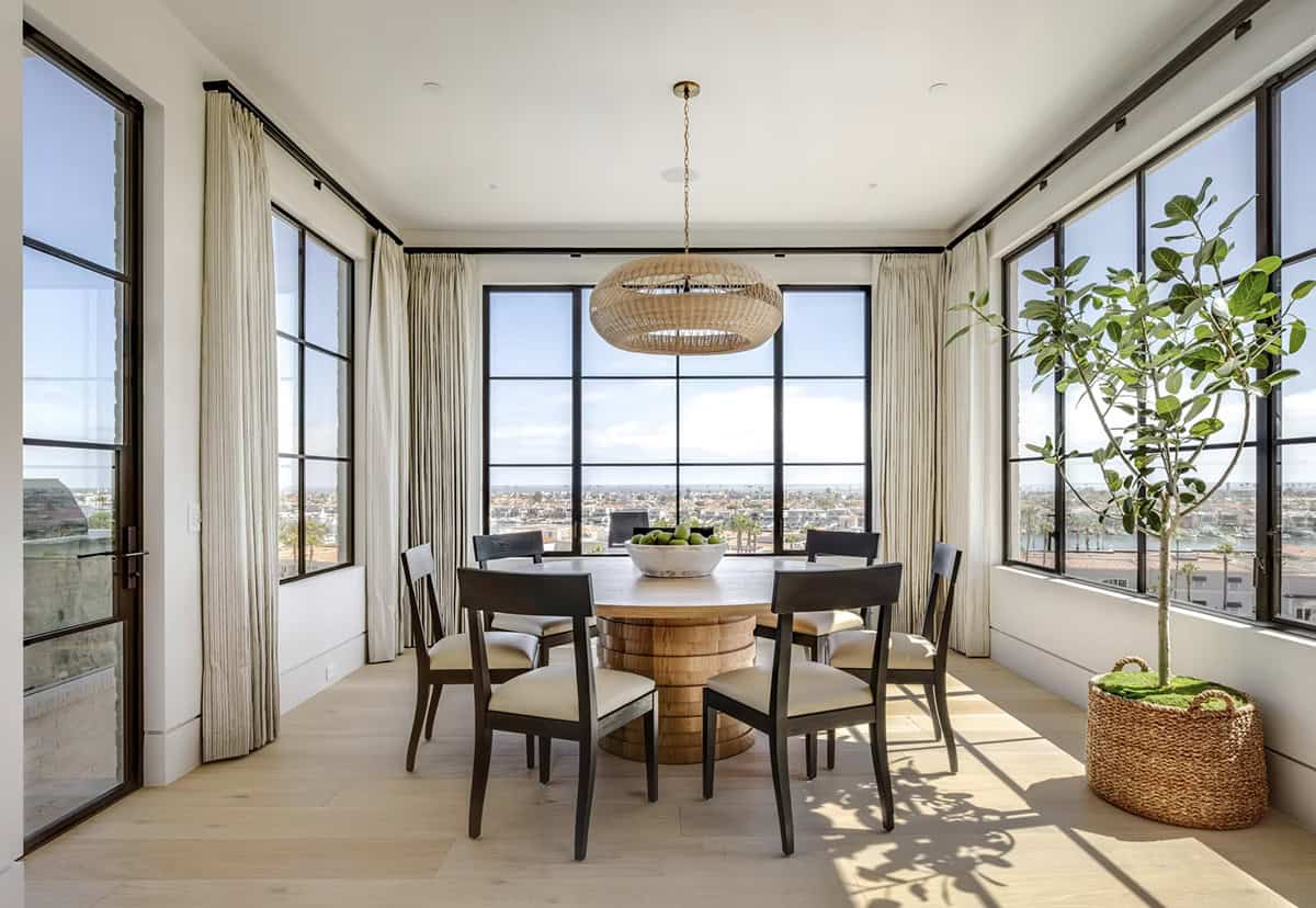 Sun-filled breakfast nook with round pedestal table, dark chairs, rattan pendant, and panoramic steel-framed windows