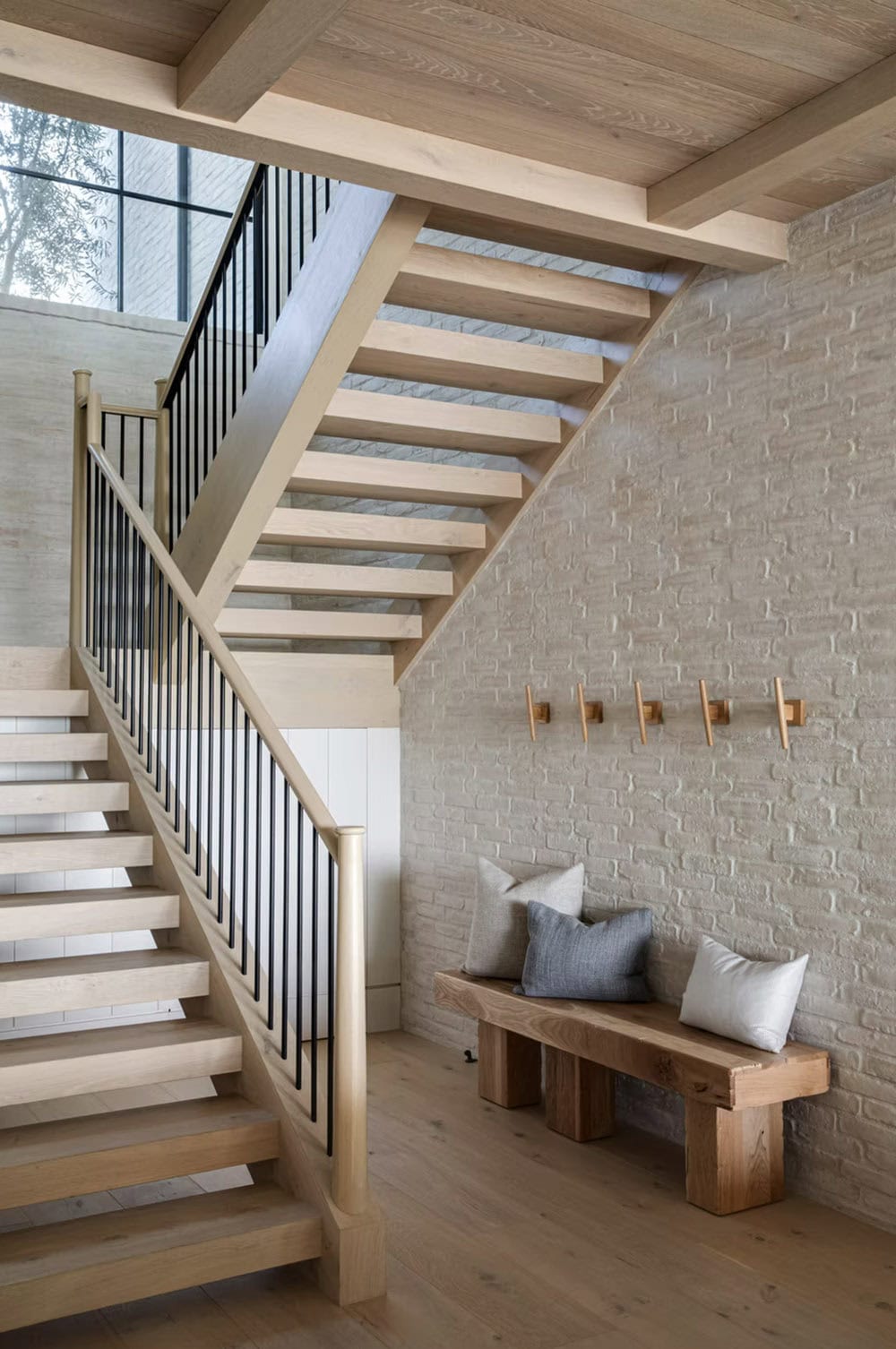 Lower-level hallway with floating oak staircase, white brick wall, mudroom bench, and brass coat hooks