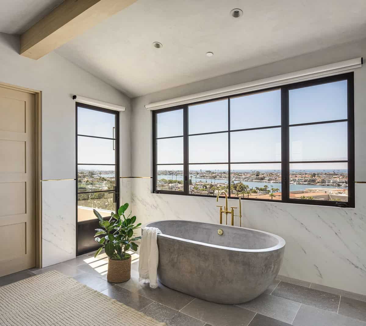 Freestanding concrete soaking tub with brass floor-mount faucet, marble surround, and panoramic harbor views