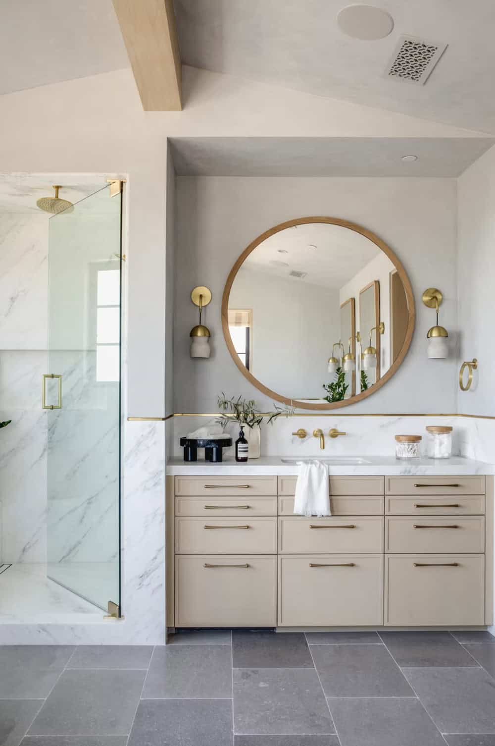 Primary bathroom vanity detail with round wood-framed mirror, brass wall sconces, and sand-colored cabinetry