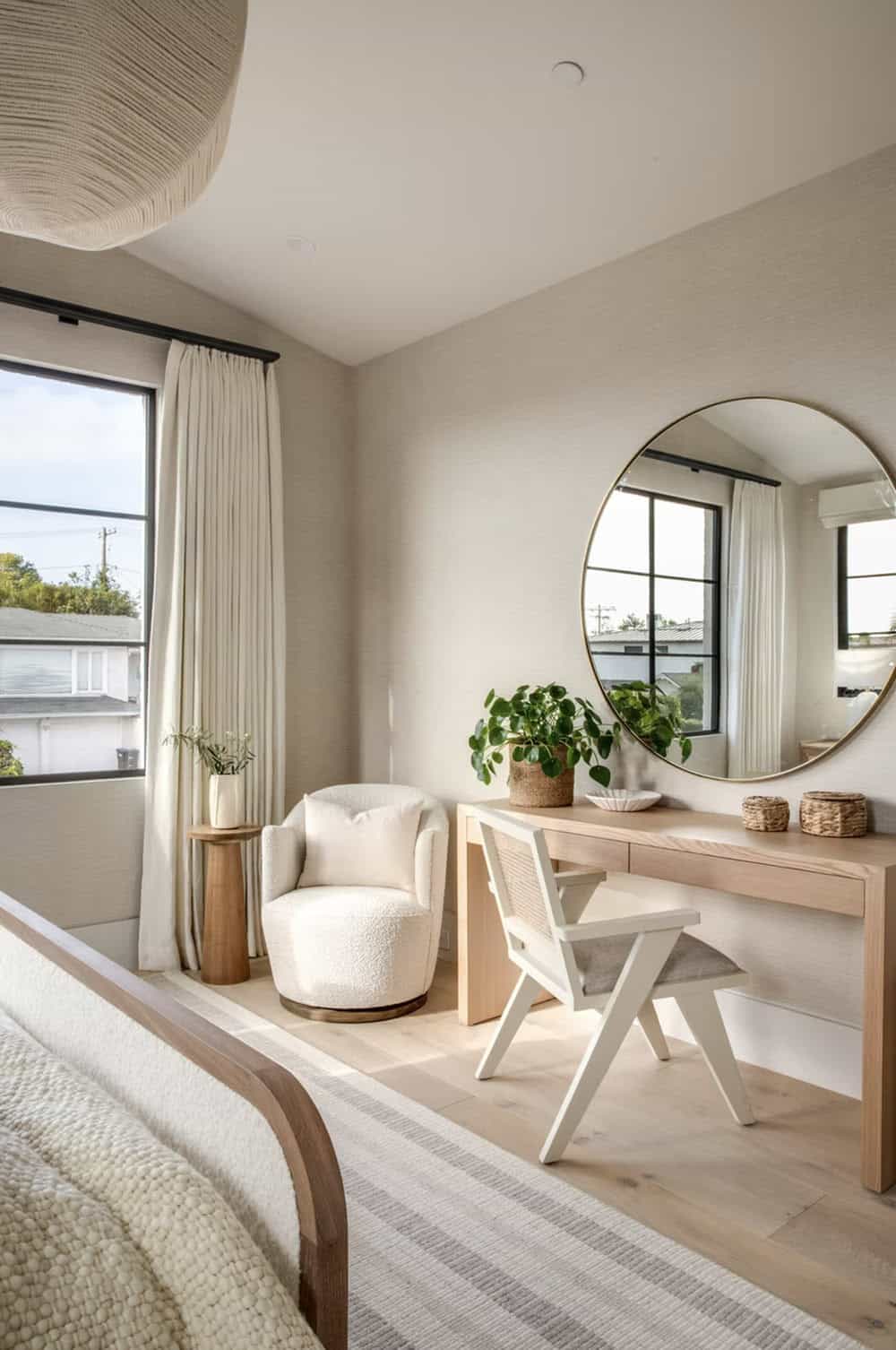 Bedroom corner with boucle swivel chair, light oak vanity desk, round brass mirror, and black-framed windows