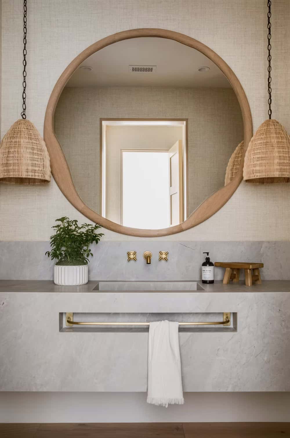 Powder room with large round wood mirror, stone floating vanity, woven pendant lights, and brass cross-handle faucet