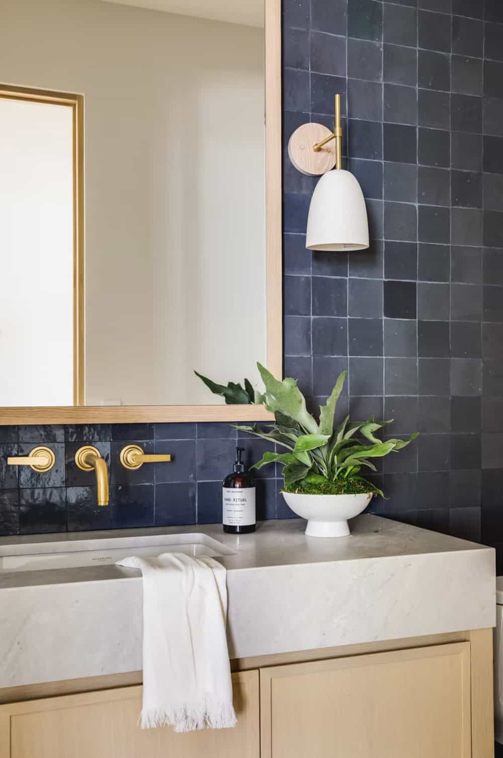 Bathroom vanity detail with dark zellige tile backsplash, marble sink, brass wall-mount faucet, and white sconce
