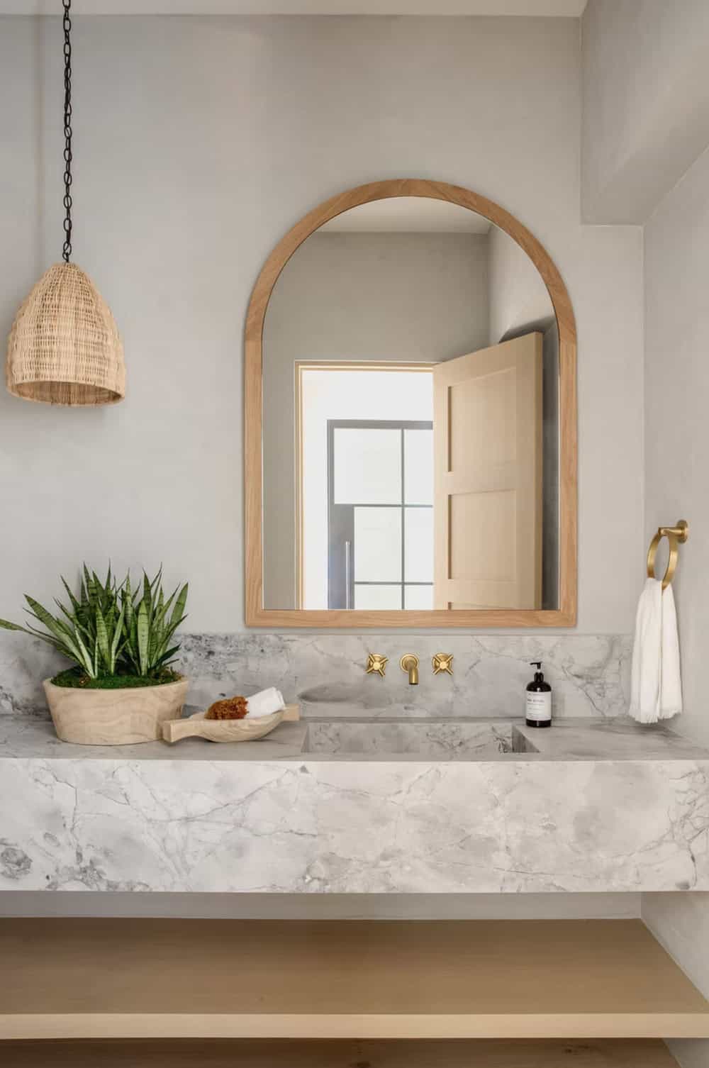 Powder room with arched wood-framed mirror, marble floating vanity, brass faucet, and rattan pendant light