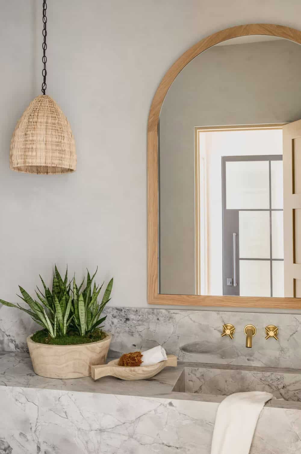Powder room close-up with arched oak mirror, marble countertop, brass cross-handle faucet, and snake plant