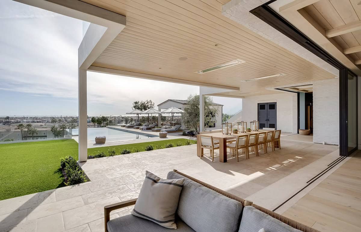 Covered outdoor living area with wood-plank ceiling, teak dining table, pool, lawn, and coastal views
