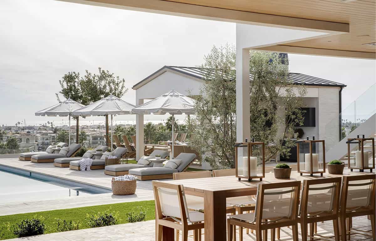 Pool deck with teak lounge chairs, striped umbrellas, outdoor kitchen, and olive trees