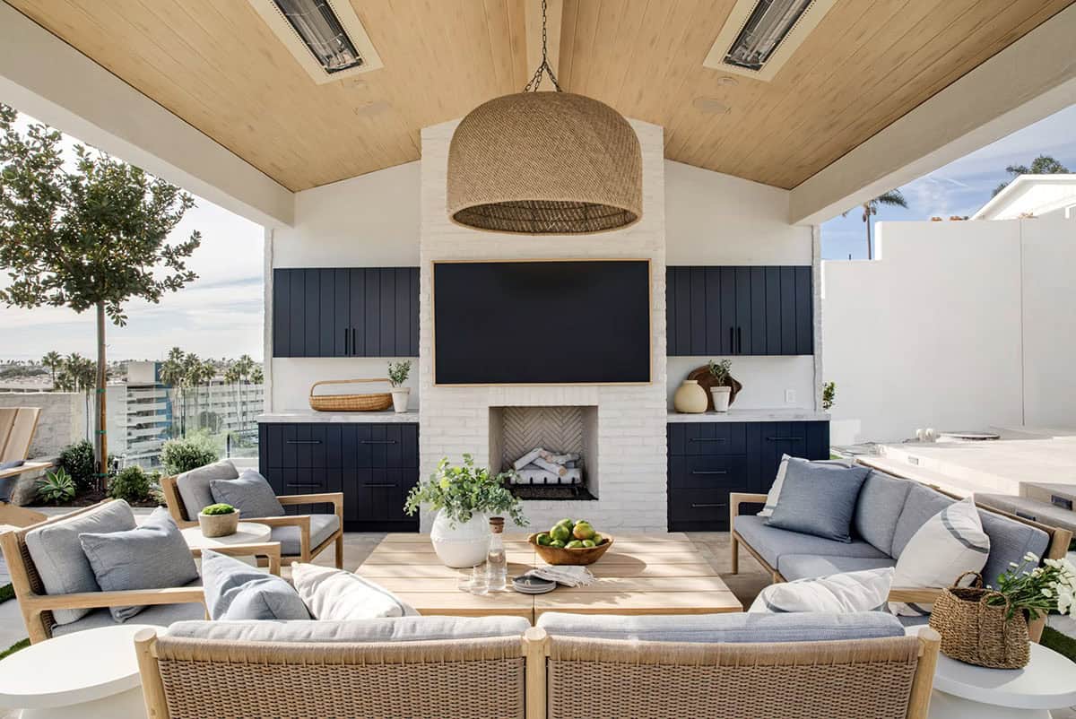 Covered outdoor living room with vaulted wood ceiling, white brick fireplace, navy cabinetry, TV, and rattan pendant