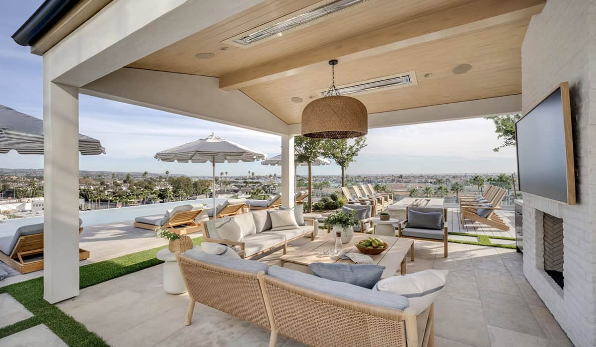 Open-air poolside lounge with rattan drum pendant, teak seating, outdoor fireplace, and coastal views