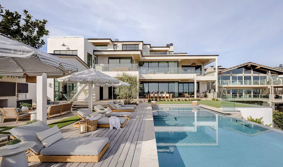 Full rear view of coastal home with infinity pool, sun deck, multiple terraces, lawn, and striped umbrellas