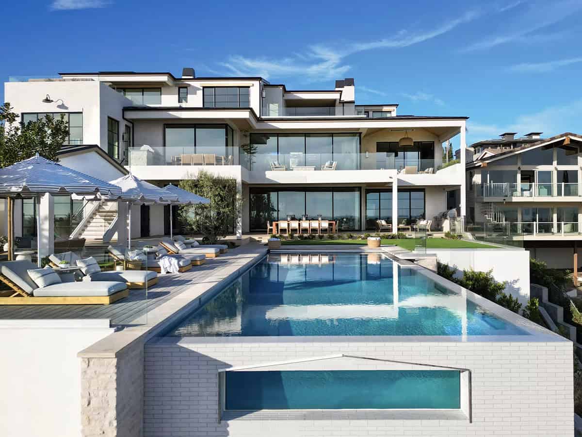 Aerial-style rear exterior showing infinity pool, glass railings, multi-level terraces, and striped pool umbrellas