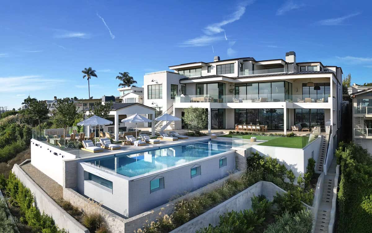 Wide aerial view of coastal contemporary home with infinity pool, terraced landscaping, and multi-story glass facade