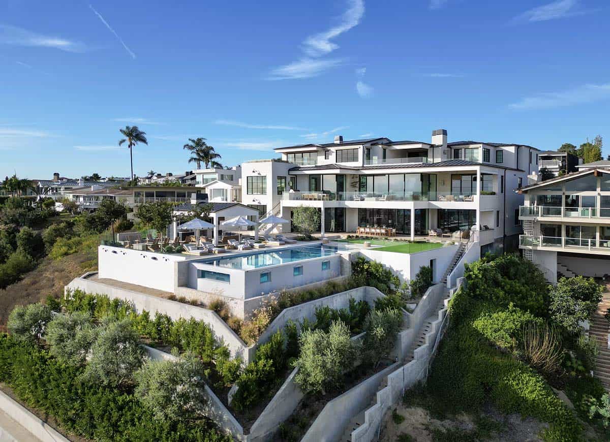 Drone view of full property showing infinity pool, terraced hillside gardens, pool deck, and white stucco exterior