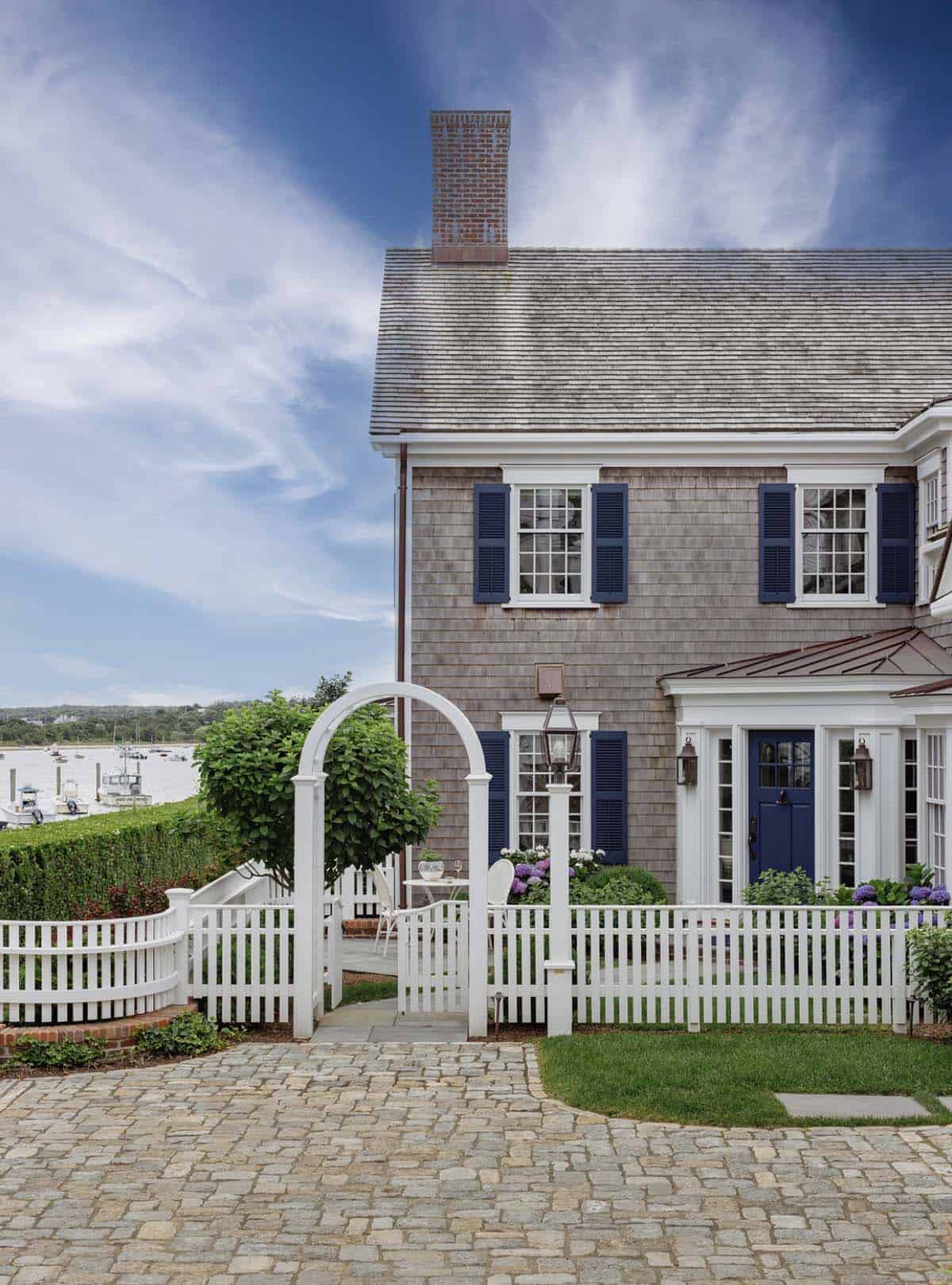 Shingle-style home exterior with navy shutters, white picket fence and arched gate