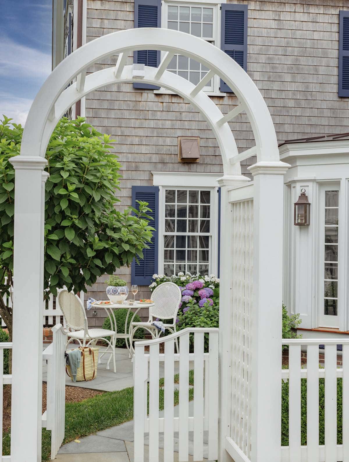 White garden arbor gate framing shingle-style home facade with hydrangeas
