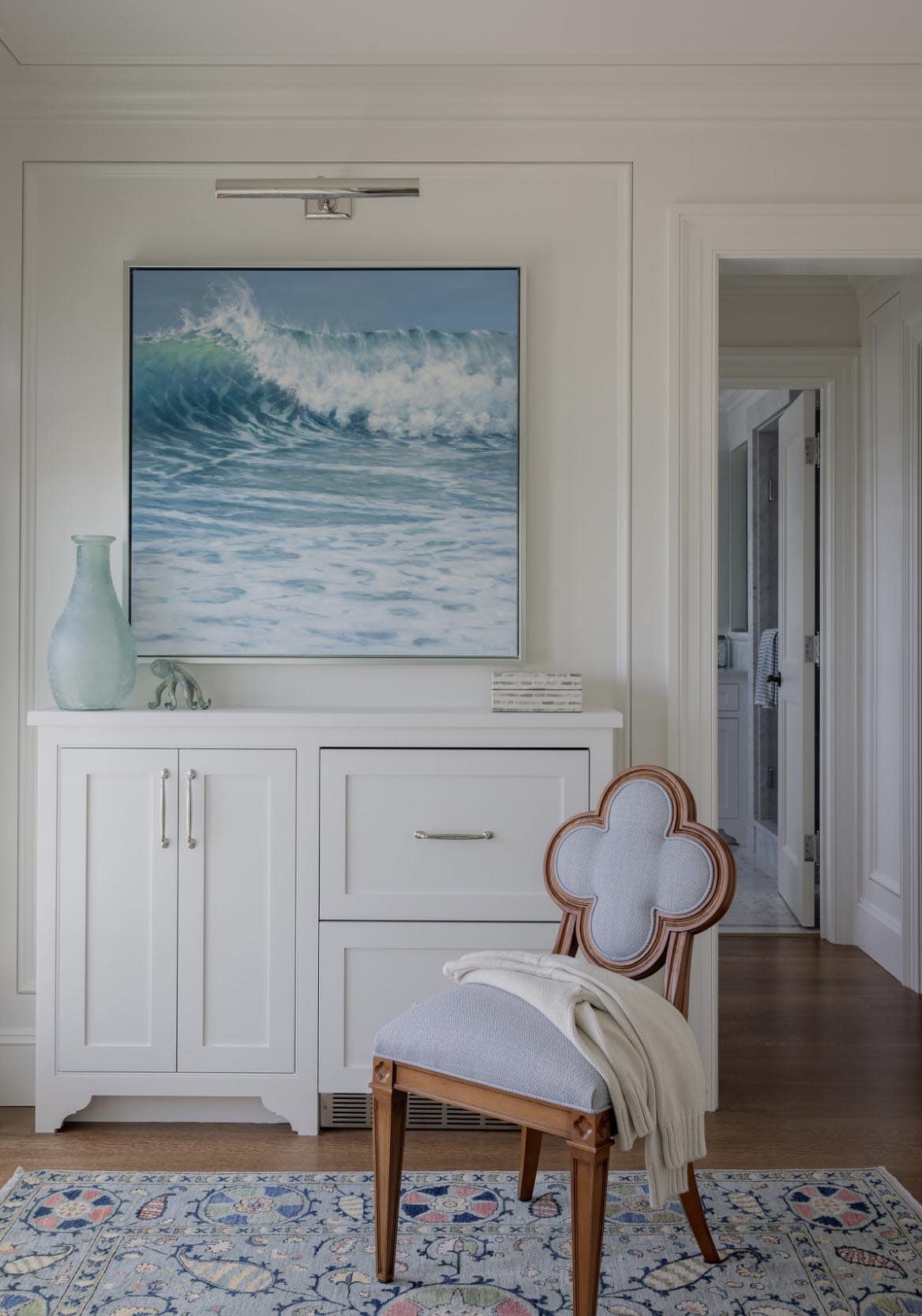 Bedroom sitting area with ocean wave painting above white built-in cabinet and quatrefoil chair