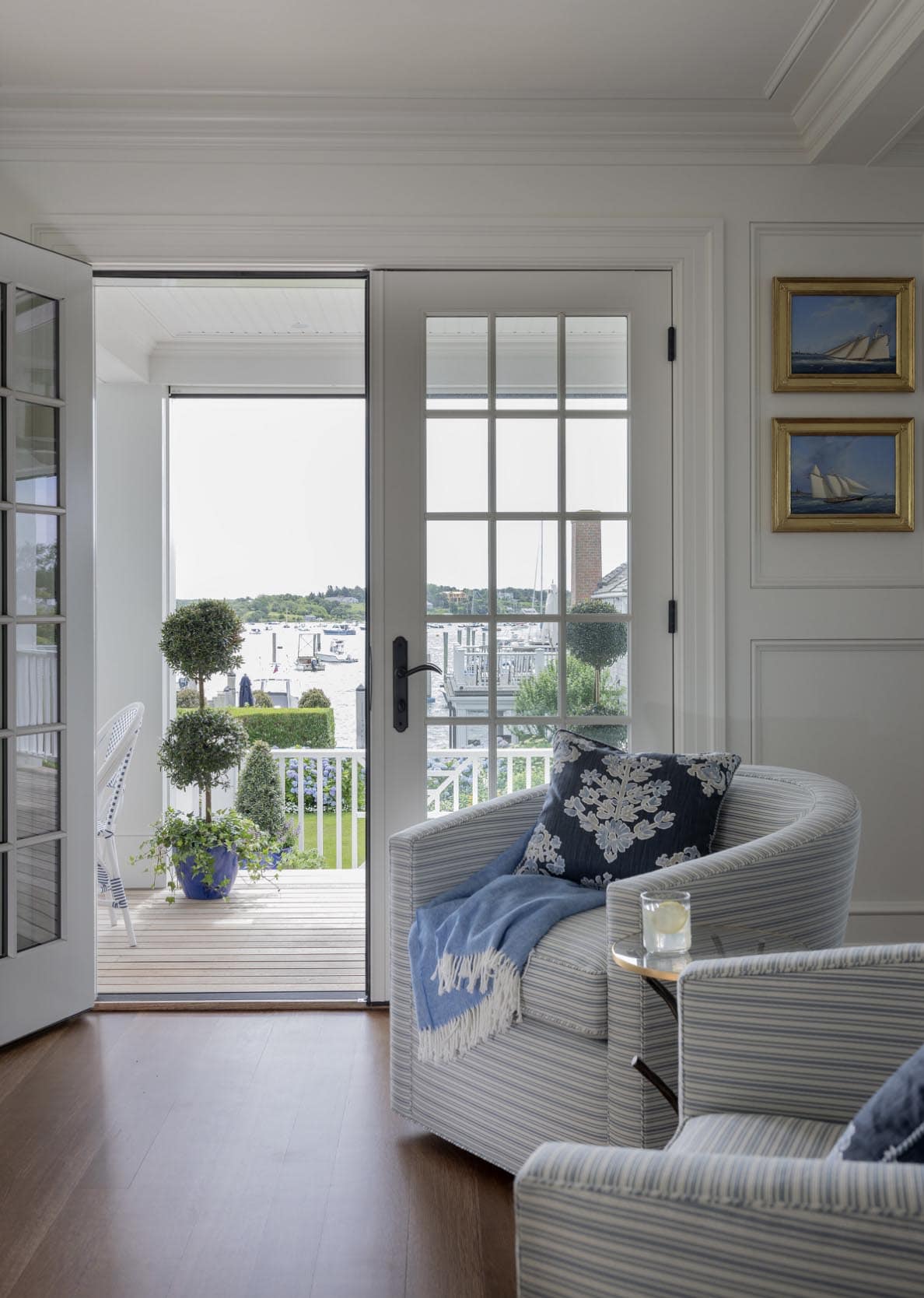 Striped armchairs beside open French doors overlooking harbor with topiary and nautical paintings