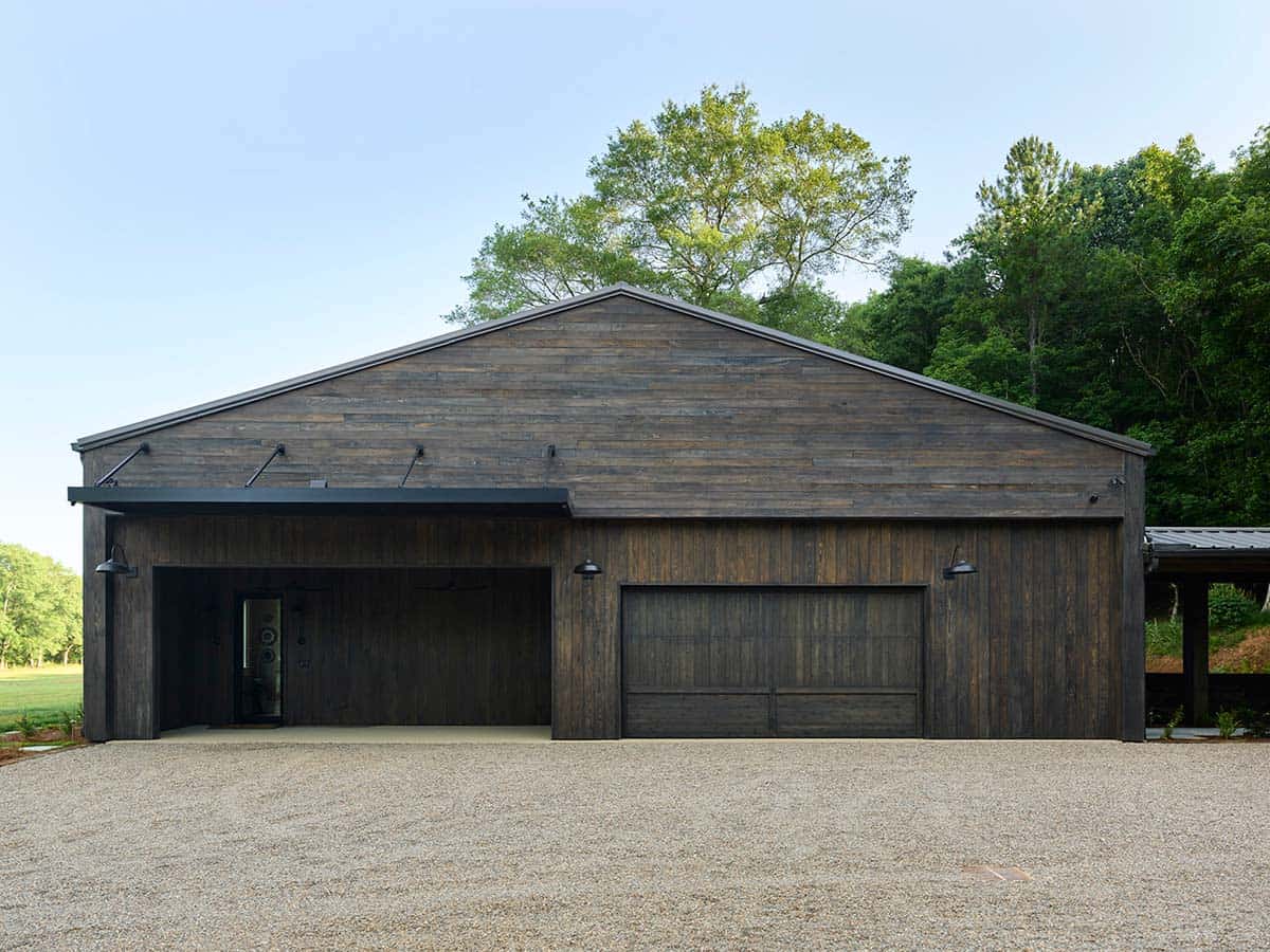 Front facade of dark weathered wood barn home with gabled roof, wood garage doors, and gravel driveway