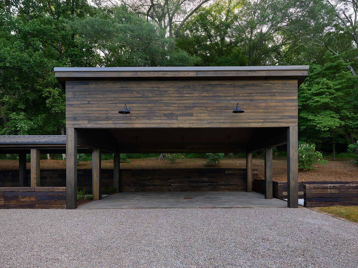 Detached open carport with weathered wood siding, flat metal roof, black barn lights, and timber columns