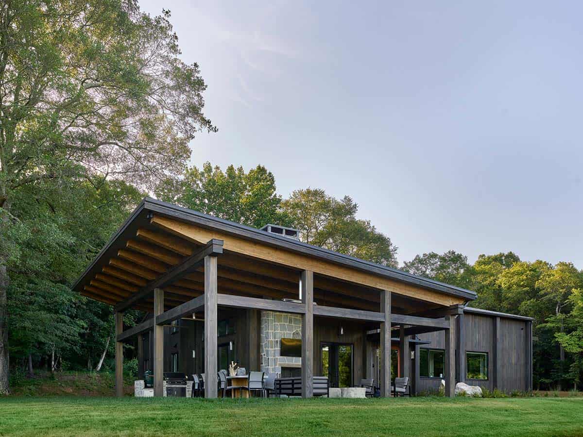 Back exterior of dark wood barn home with expansive timber roof overhang, stone fireplace, and outdoor living