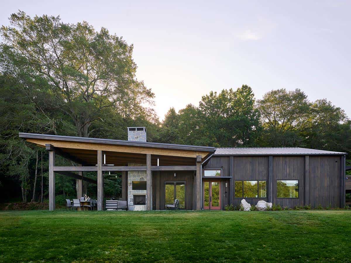 Dusk view of contemporary barn home showing covered porch, stone chimney, red entry doors, and green lawn