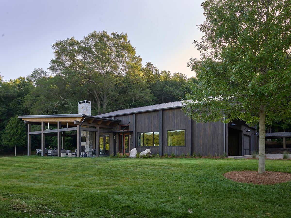 Evening exterior of dark wood contemporary home with covered patio, stone fireplace chimney, and wooded backdrop