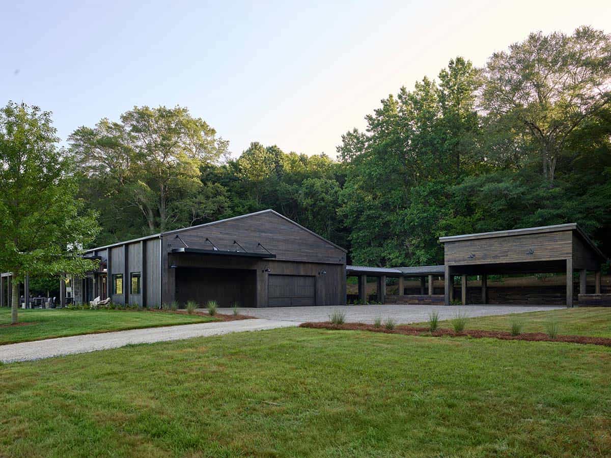 Wide view of barn-style home with detached carport structure, gravel motor court, and mature tree-lined setting